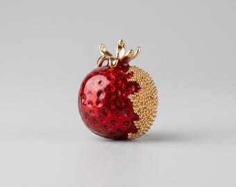 May include: A red and gold strawberry-shaped brooch. The brooch has a glossy red body with a textured surface and a section of gold-colored beads. The top features a gold-colored crown-like design.