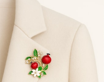 May include: A gold-tone metal brooch featuring two red pomegranate fruits with textured surfaces, green enameled leaves, a white five-petaled flower, and small clear crystals attached to the branches, pinned to a cream-colored blazer.