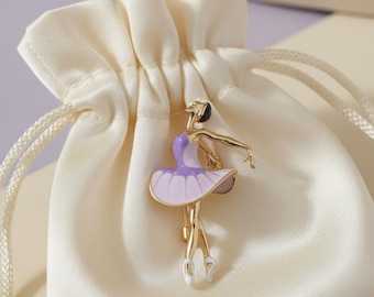 May include: A gold-colored ballerina brooch with a purple and lavender tutu, poised in a graceful dance. The brooch is set against a cream-colored satin pouch with a drawstring closure, suggesting a gift or keepsake.