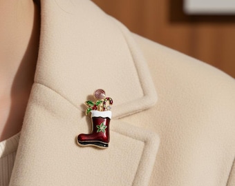 May include: A Christmas-themed brooch in the shape of a red and black boot, adorned with holly, candy canes, and a swirl. The brooch is pinned to a beige blazer, suggesting festive holiday attire.