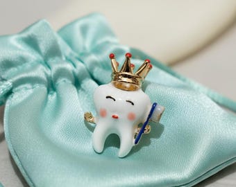 May include: A white tooth-shaped brooch with a gold crown and a small blue toothbrush. The brooch has a cute, cartoonish design with rosy cheeks and closed eyes. It rests on a light blue satin drawstring bag.