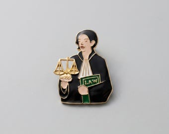 May include: Enamel pin depicting a person in a black robe holding a golden scale and a green book with the word "LAW". The pin has a white border and is set against a gray background.
