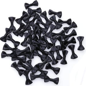 May include: A collection of small, black satin bow ties scattered on a white surface. The bows are uniform in size and shape, with a classic bow tie design. The satin material gives them a slight sheen.