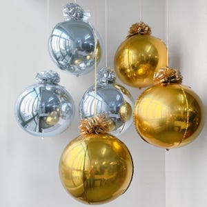 May include: A collection of six spherical balloons in metallic gold and silver finishes, each topped with a matching decorative ribbon bow. The balloons are suspended at varying heights, featuring highly reflective surfaces that mirror the room.