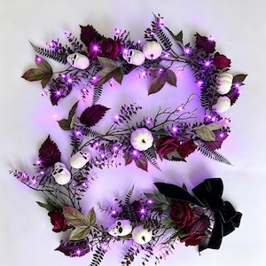 May include: A decorative garland featuring dark burgundy roses, green and dark purple foliage, white miniature pumpkins, and small white skulls. The arrangement is illuminated by bright purple LED fairy lights and finished with a black velvet bow.