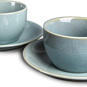 May include: Two matching blue-grey ceramic teacups and saucers. The cups have a rounded shape with a handle, and the saucers are circular. The rims of the cups and saucers have a light beige trim. The set is designed for serving tea or coffee.