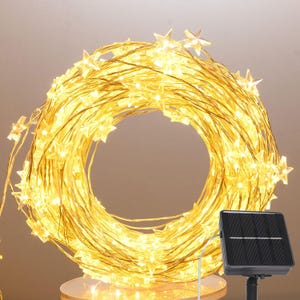 May include: A coiled string of warm white LED fairy lights featuring small star-shaped covers. The lights are connected to a black rectangular solar panel on a stake, designed to power the illumination through solar energy collection.