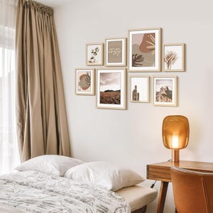 May include: A collection of framed art prints in a bedroom setting. The prints feature neutral tones and abstract designs, including a landscape and the text "let's get cozy". The frames are light wood, and the arrangement is above a bed.