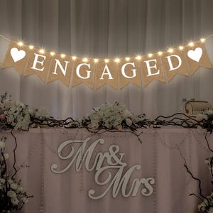 May include: A burlap banner with white letters spelling "ENGAGED" and white heart shapes on each end. The banner is strung with white lights and hangs above a table decorated with white flowers and a sign that says "Mr. & Mrs."