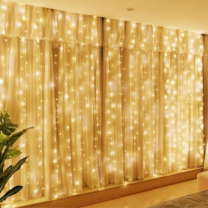 May include: A set of sheer, cream-colored curtains illuminated by warm white fairy lights. The lights are evenly spaced, creating a soft, glowing effect. The curtains are hanging in front of a window, creating a cozy ambiance.