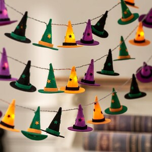 May include: A string of LED fairy lights featuring miniature felt witch hats in orange, purple, green, and black. Each hat is decorated with small star cutouts, and the lights are integrated within the hats to provide a warm glow.