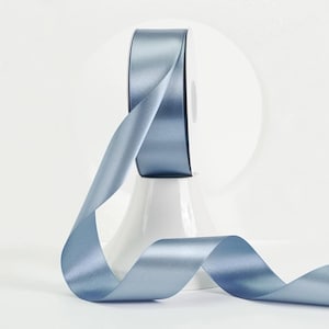 May include: A spool of light blue satin ribbon. The ribbon is unwound, with a portion of the ribbon still on the spool. The ribbon is displayed on a white, decorative stand. The ribbon has a smooth, shiny texture.