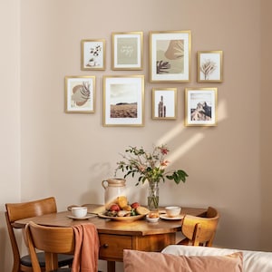 May include: A gallery wall arrangement of nine framed art prints with gold frames. The prints feature abstract designs, landscapes, and botanical illustrations. The word "cozy" is visible in one of the prints. The wall is painted a neutral color.