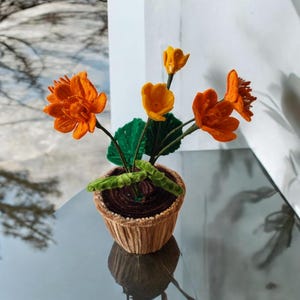 May include: A small potted plant with orange and yellow felt flowers and green felt leaves. The pot is brown and has a ribbed texture.