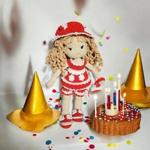 Könnte beinhalten: Eine gehäkelte Puppe, die einen rot-weißen Badeanzug und einen roten Hut mit einer Blume trägt. Die Puppe steht neben einem Geburtstagskuchen mit brennenden Kerzen.