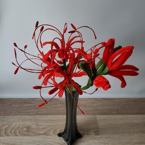 May include: A striking arrangement of red flowers with delicate, elongated petals and stamens, displayed in a tall, black vase. The flowers have a textured look, and the vase has a flared base. The arrangement is on a wooden surface.