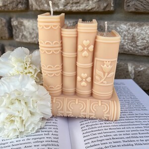 May include: A set of five stacked book-shaped candles in a peach colour. The candles have a decorative design of flowers and swirls. The candles are sitting on a book.