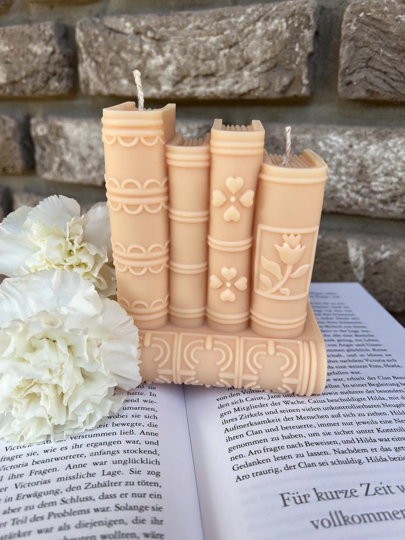 May include: A set of five stacked book-shaped candles in a peach colour. The candles have a decorative design of flowers and swirls. The candles are sitting on a book.