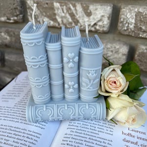 May include: Three blue book-shaped candles with white wicks and decorative details. The candles are stacked on top of each other and are surrounded by white roses.