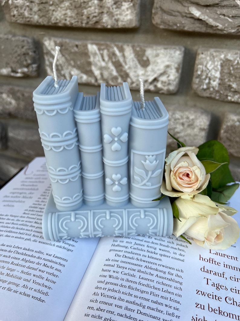 May include: Three blue book-shaped candles with white wicks and decorative details. The candles are stacked on top of each other and are surrounded by white roses.