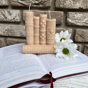May include: A set of three beige pillar candles shaped like books with decorative designs. The candles are sitting on top of a book with a white daisy flower on top.