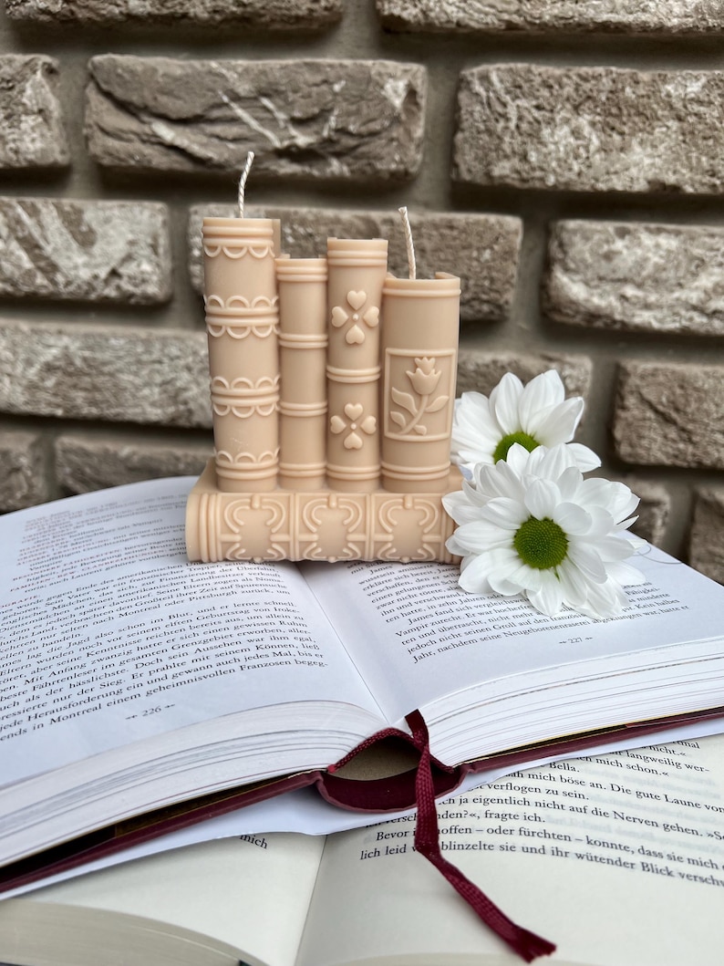 May include: A set of three beige pillar candles shaped like books with decorative designs. The candles are sitting on top of a book with a white daisy flower on top.