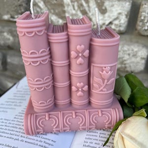 May include: A set of four pink book-shaped candles with decorative details. The candles are stacked on top of each other and have a white wick.