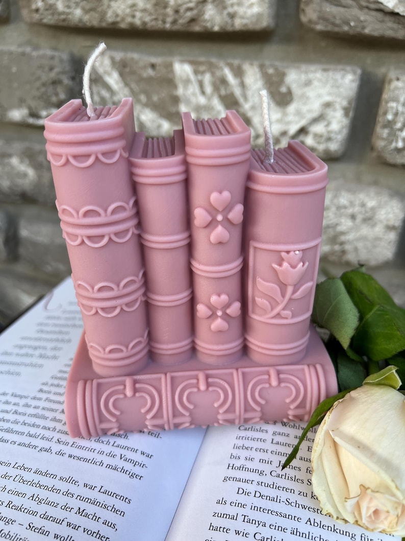 May include: A set of four pink book-shaped candles with decorative details. The candles are stacked on top of each other and have a white wick.