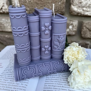 May include: A set of four purple book-shaped candles with white wicks. The candles are stacked on top of each other and have decorative details on the spines. The candles are sitting on a book with white flowers in the background.
