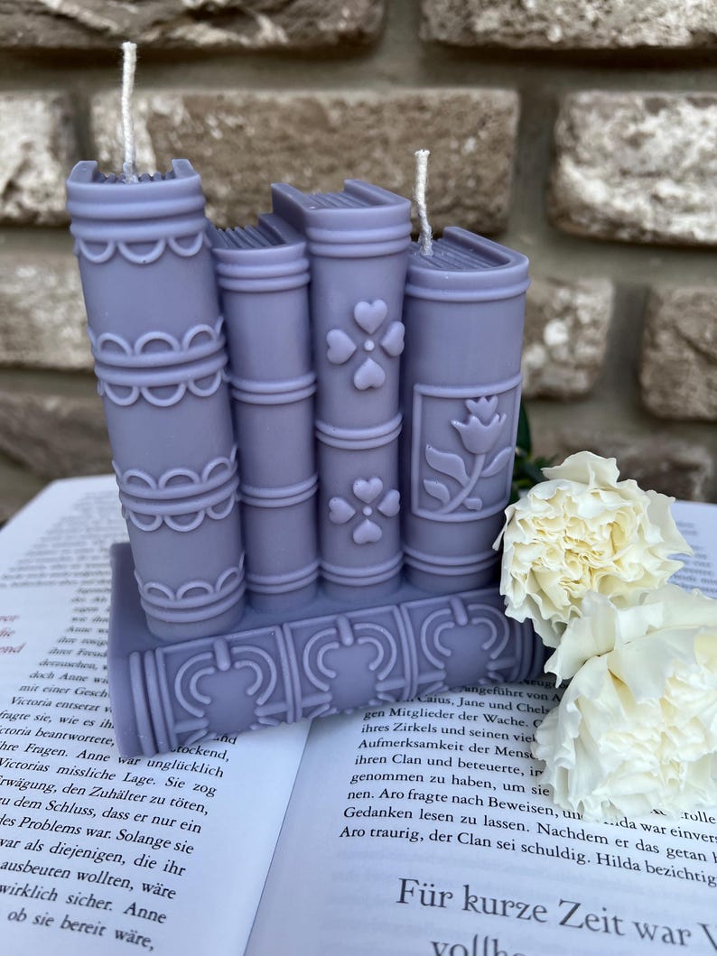 May include: A set of four purple book-shaped candles with white wicks. The candles are stacked on top of each other and have decorative details on the spines. The candles are sitting on a book with white flowers in the background.