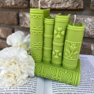 May include: A set of five green book-shaped candles with decorative details. The candles are arranged in a stack, with the largest candle at the bottom and the smallest candle at the top. The candles are made of a smooth, waxy material and have a slightly textured surface.