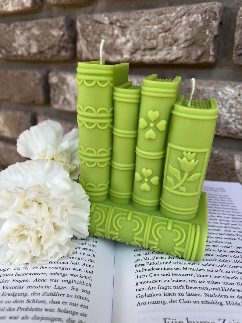 May include: A set of five green book-shaped candles with decorative details. The candles are arranged in a stack, with the largest candle at the bottom and the smallest candle at the top. The candles are made of a smooth, waxy material and have a slightly textured surface.