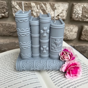 May include: A stack of four grey book-shaped candles with intricate designs. The candles are arranged on a book with a white background. The candles have a white wick and are decorated with a floral pattern.