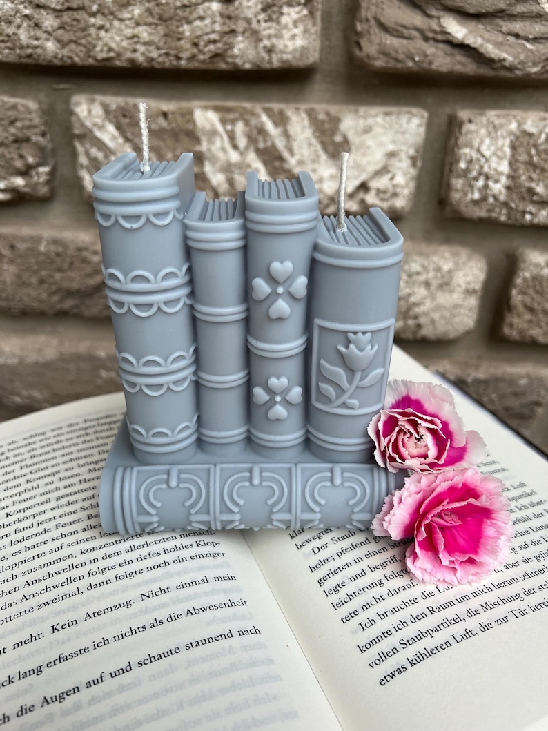 May include: A stack of four grey book-shaped candles with intricate designs. The candles are arranged on a book with a white background. The candles have a white wick and are decorated with a floral pattern.