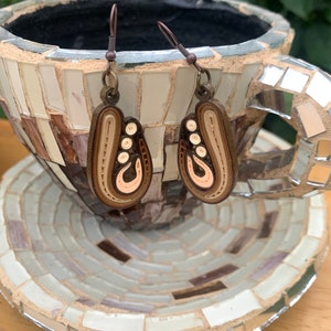 May include: A pair of brown and cream paper quilling earrings with a swirl design. The earrings are hanging from a brown metal hook.
