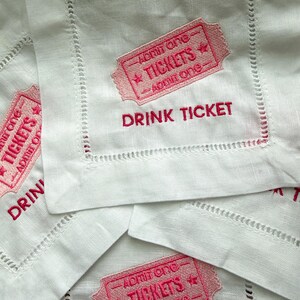 Puede incluir: Posavasos de lino blanco con dise&ntilde;os de boletos bordados en rosa y las palabras "DRINK TICKET". Cada boleto presenta el texto "ADMIT ONE" y "TICKETS". Los posavasos tienen un borde cosido.