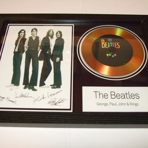 May include: A framed gold disc award featuring a black and white photograph of The Beatles. The photo is signed by all four members. The disc is inscribed with "The Beatles" and "George, Paul, John & Ringo".