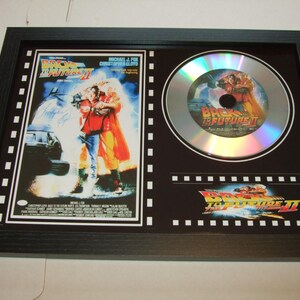 May include: A framed silver CD with a black and white movie poster of Back to the Future Part II. The poster features Michael J. Fox and Christopher Lloyd. The CD has a silver background with a black and white image of the DeLorean time machine.