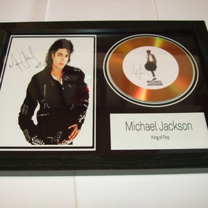 Puede incluir: Exhibición enmarcada con una fotografía y un disco de oro. La fotografía muestra a un hombre con una chaqueta negra. El disco de oro es circular con una silueta y firma. El texto dice "Michael Jackson King of Pop".