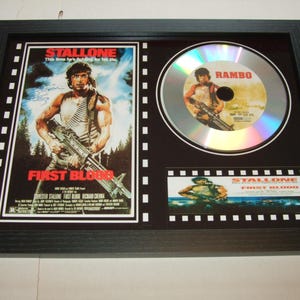 Puede incluir: Un expositor de póster de película y CD enmarcado en negro con Sylvester Stallone en la película "First Blood". El póster presenta el título "First Blood" y el texto "Esta vez está luchando por su vida". El CD es un disco plateado con el título "Rambo" impreso en él.