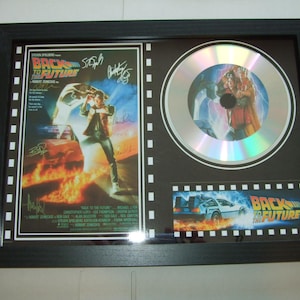 Puede incluir: Una pantalla enmarcada en negro que presenta un disco plateado con una imagen holográfica del DeLorean, la máquina del tiempo de la película Regreso al futuro. La pantalla también incluye un póster de película con el título "Regreso al futuro" y varias firmas.