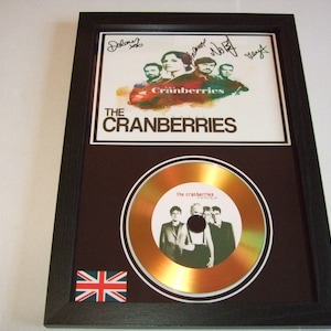 May include: A framed gold disc featuring a picture of the band The Cranberries. The disc is signed by the band members. The frame is black with a Union Jack flag in the bottom right corner.