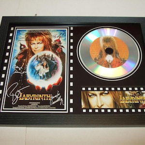May include: A black framed display with a silver disc featuring a character from the movie Labyrinth. The disc is surrounded by a black mat with a film strip border. The display also features a poster of the movie Labyrinth with a signature from the actor David Bowie. The poster is also surrounded by a black mat with a film strip border.