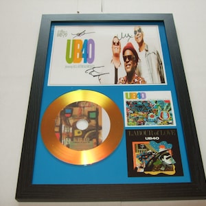 Puede incluir: Un disco de oro enmarcado con fondo azul. El disco es un disco de oro con un diseño colorido. El marco incluye una foto de la banda UB40 con sus firmas. La portada del álbum "Labour of Love" también está incluida en el marco.