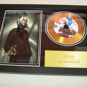 以下が含まれることがあります： ポール・ウェラーの白黒写真が飾られた額縁に入ったゴールドレコード。レコードはゴールドで、赤、白、青のデザインが施されています。レコードの下のプレートには「ポール・ウェラー 1962年5月25日生まれ、ウォーキング、サリー」と書いてあります。