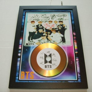 Puede incluir: Un disco de oro enmarcado con una foto del grupo de K-pop BTS. El disco está rodeado por un diseño de galaxia en blanco y negro. El disco tiene el nombre de la banda "BTS" impreso en él. La foto muestra a los miembros de la banda sosteniendo un cartel en forma de corazón que dice "Te amo". La foto está firmada por los siete miembros de la banda.