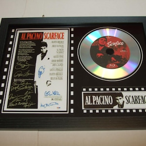 Puede incluir: Un marco negro que presenta un disco Blu-ray plateado con un logotipo "Scarface" rojo y blanco. El disco está rodeado por una cenefa negra con un borde de tira de película blanca. El lado izquierdo del marco presenta una foto en blanco y negro de Al Pacino como Tony Montana de la película "Scarface" con el título "Al Pacino Scarface" impreso sobre la foto. La foto está rodeada por un borde blanco con bordes de tira de película negros. El texto "Al Pacino Scarface" está impreso en blanco sobre una tira de película negra en la parte inferior del marco.