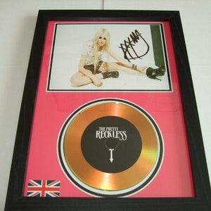 May include: A framed gold record award with a black and white image of a woman in a white shirt and black boots. The record is for "The Pretty Reckless" and features a black and white image of a downward pointing arrow with a cross on top. The frame has a pink background and a Union Jack flag in the bottom left corner.