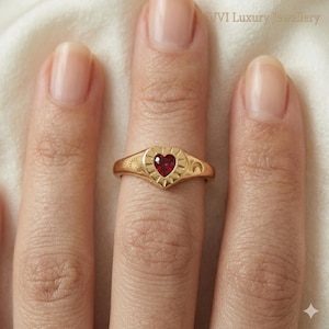 May include: A gold ring with a heart-shaped red gemstone. The ring has sun and moon designs on the sides. The ring is on a finger.
