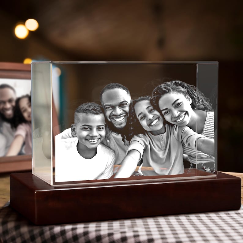 Puede incluir: Un bloque de fotos rectangular transparente que muestra un retrato familiar en blanco y negro. El bloque se asienta sobre una base de madera oscura. Otra foto enmarcada es visible en el fondo. La familia incluye dos ni&ntilde;os y dos adultos, todos sonriendo.